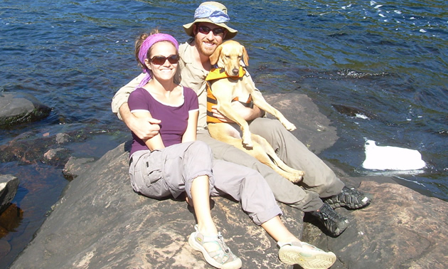 Smiling young couple and their big golden lab dog sit on a large rock that extends into a body of water