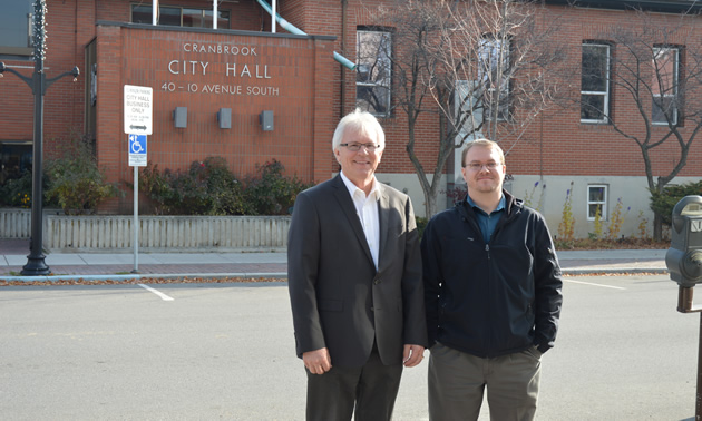 Two men standing with red brick building—Cranbrook City Hall—in the background