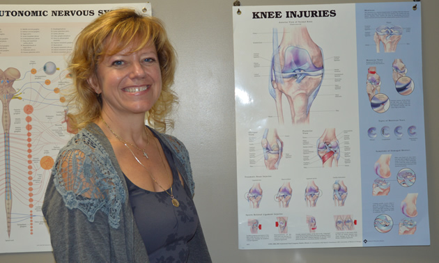 Close-up of a smiling woman with blue eyes and blonde hair, with anatomical charts on the wall behind her