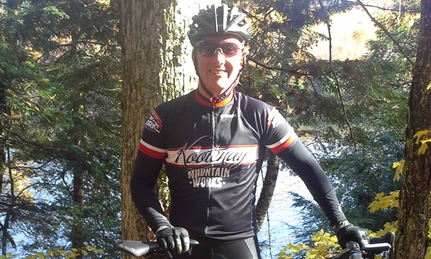 Smiling man in outdoor autumn setting, wearing a cycling helmet and jacket with a logo across the chest, and holding onto a bicycle.