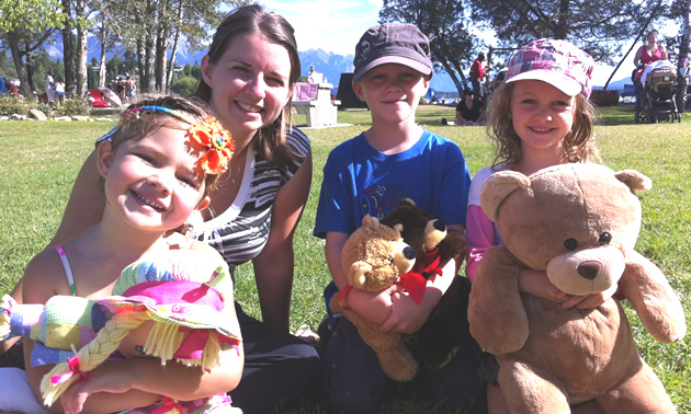 Young woman and three children with teddybears sit on the grass in a park