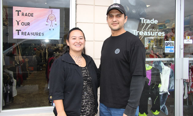 Young middle-aged couple stand in front of a store displaying Trade Your Treasures sign.