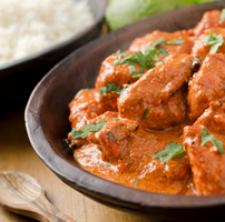 A bowl of spicy red butter chicken is sprinkled with cilantro. Rice, limes and a wooden spoon are in the background.