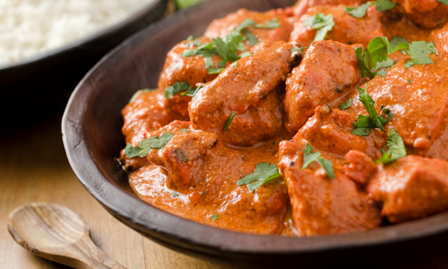 A bowl of spicy red butter chicken is sprinkled with cilantro. Rice, limes and a wooden spoon are in the background.