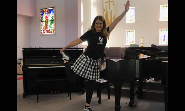 Tanya DuBois is standing in front of a piano, stretching one leg behind her and raising one arm in the air.
