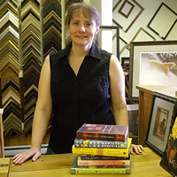 Trish Bazalgette of Tara Books in Salmo stands behind a counter with frames on the wall and a stack of books.