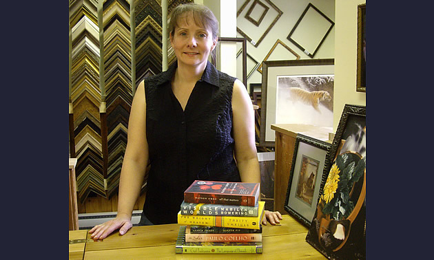 Trish Bazalgette of Tara Books in Salmo stands behind a counter with frames on the wall and a stack of books.
