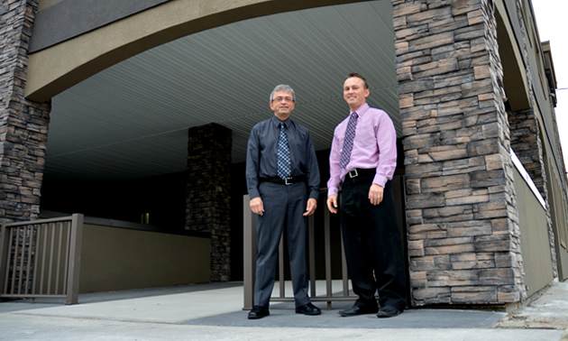 Two men in shirtsleeves and ties stand next to a pillar of stone and a finished arch.