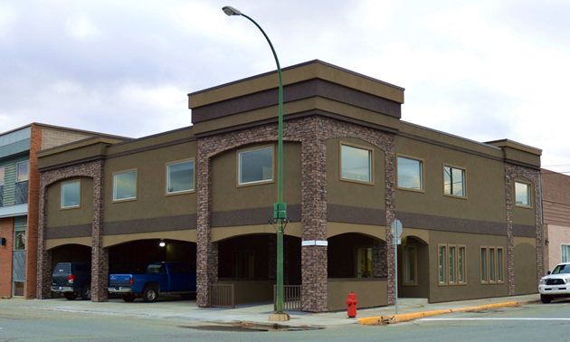 A building in shades of grey/green with stone pillars and trim stands on the corner of a street.