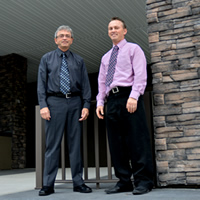 Two men in shirtsleeves and ties stand next to a pillar of stone and a finished arch.