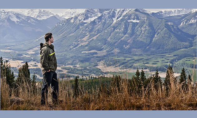 A man stands overlooking a valley with mountains in the background.