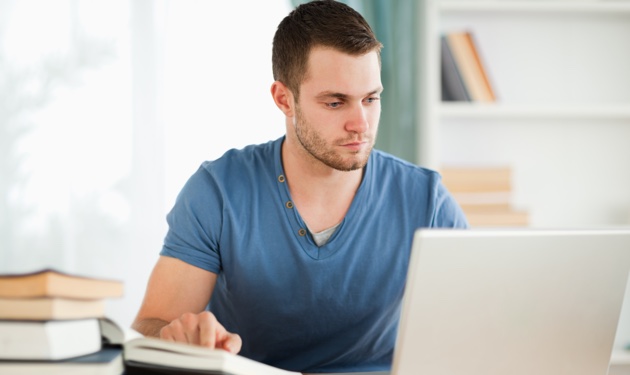 A young man sitting in front of a laptop with a stack of books beside him.