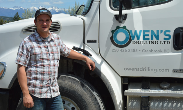 Young man in jeans, summer shirt and baseball cap stands beside a big white truck with Owen's Drilling on the driver door