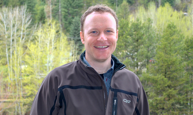 Tim Hicks is standing in the outdoors, wearing a gear jacket and smiling for the camera.