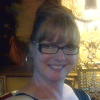 Tracy Floer has red/blonde hair in an updo, and glasses. She stands in front of an antique lamp.