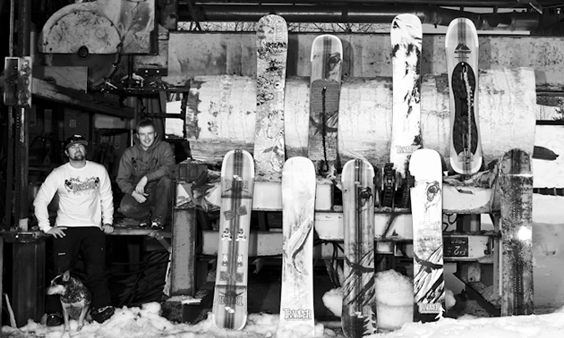 Riddell and Fortier couch alongside a display of their boards which are placed in an industrial setting. Photo is black and white.