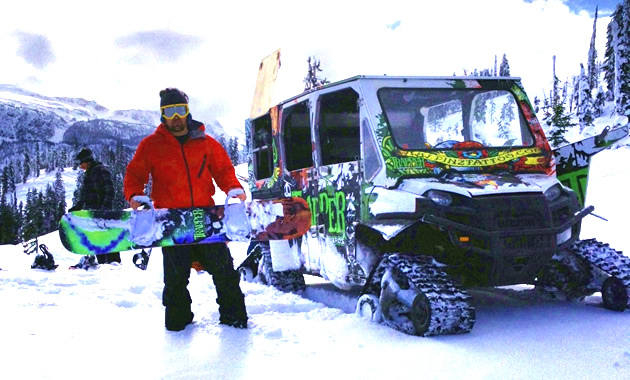 Tyler, wearing a red jacket, holds a bright snowboard against a brighter snowcat.