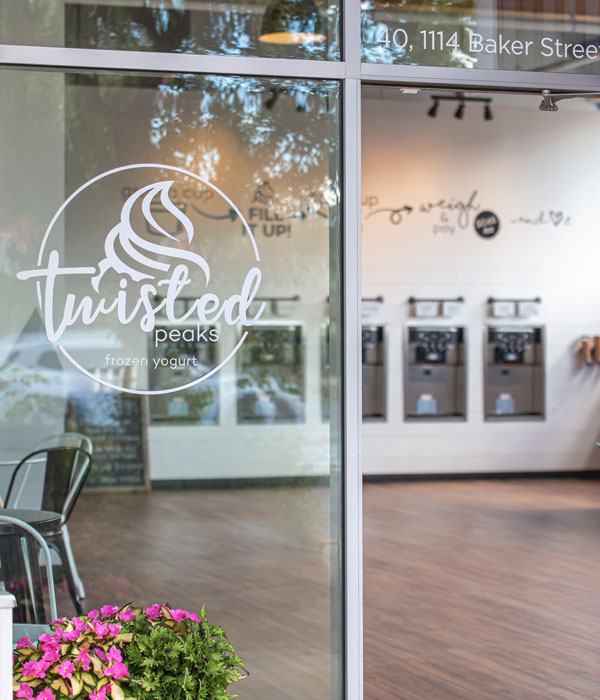 Outside of Twisted Peaks shop, showing logo on glass windows and row of yogurt machines in distance. 