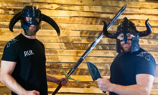 Two people, owners of Ullr's Bar, are wearing Viking hats and holding swords. 