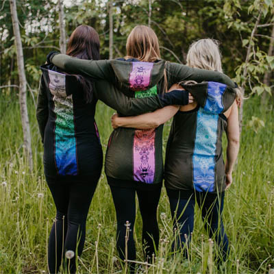 Three women wearing long black hoodies with colourful back panels, standing with backs to camera in wooded area. 