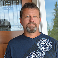 Bearded man in a blue T-shirt stands at the door of a house.