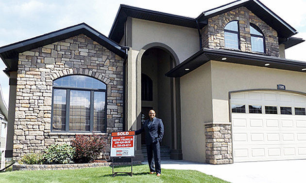 Wayne Franklin holding on to a for sale sign outside a large brick home. 