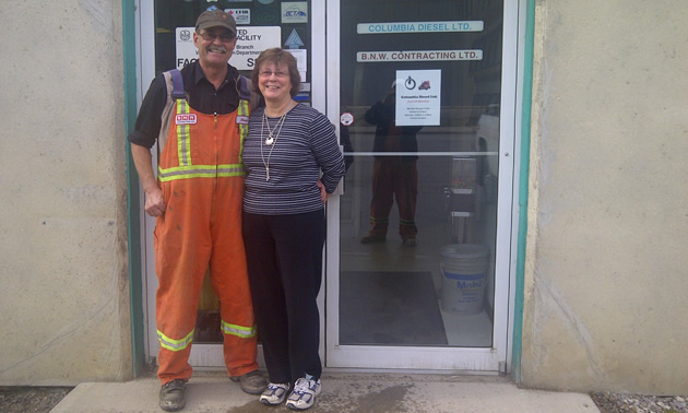 Smiling senior couple stand in front of glass doors bearing the sign Columbia Diesel Ltd. and BNW Contracting Ltd.