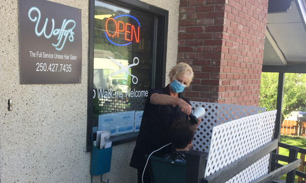 Hairdresser outside on shop deck cutting a customer's hair. 