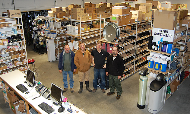 The camera looks down on four men standing in a shop surrounded by water-service parts.