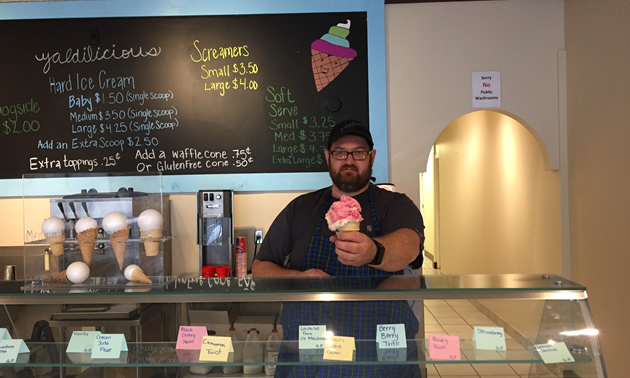 Kendyn Mackenzie, bearded and wearing a baseball hat, stands behind the glass showcase in his ice cream shop, offering a huge pink ice cream cone