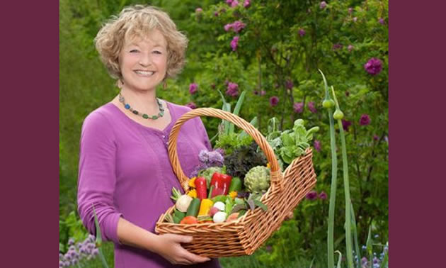 A women wears a pink cardigan and holds a basket overflowing with produce.