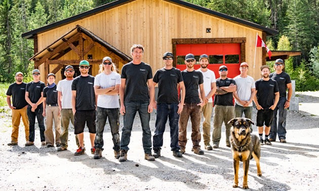 A large crew of men and one dog are lined up in a large vee in front of a shop. Rexxy, the dog, is the most distracting crew member. He shows true professionalism when welcoming people to the shop.