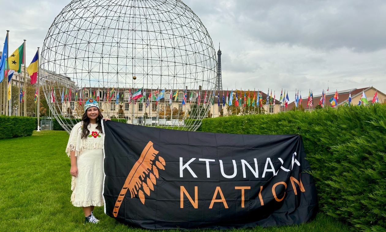 Aiyana in traditional dress, standing beside a Ktunaxa Nations sign in front of the United Nations building