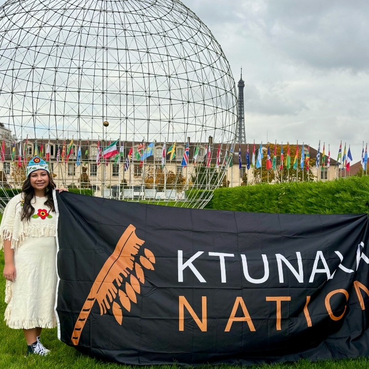 Aiyana in traditional dress, standing beside a Ktunaxa Nations sign in front of the United Nations building