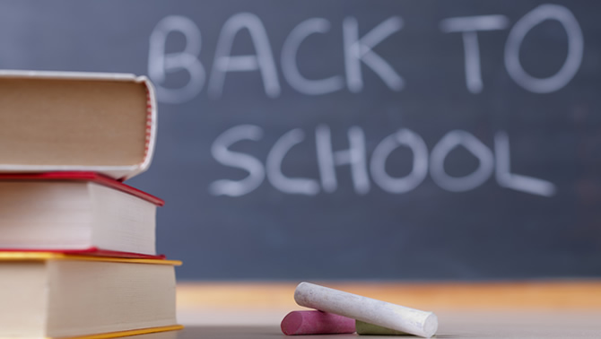 Picture of chalk and books sitting on desk, with the words 'Back to School' written on a blackboard in the background. 