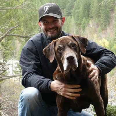 A middle-aged man standing beside a brown chocolate lab. 
