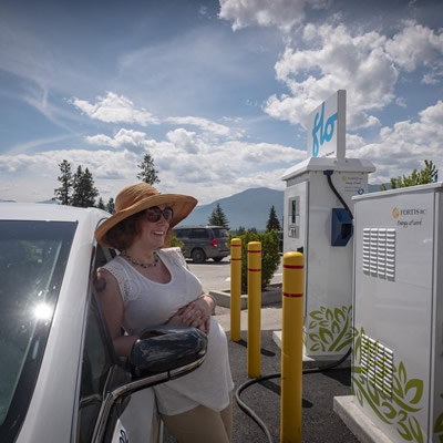 A woman with a hat leans on her car at a EV-charging station. 