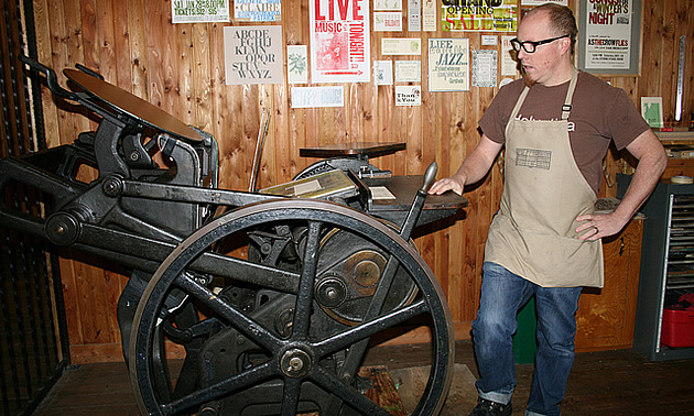 Man standing beside an old-fashioned letterpress machine in Fernie, BC