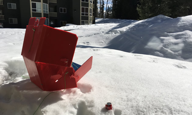 A red solar cooker is sitting in the snow outside an apartment building on a sunny winter day.