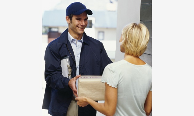 A delivery man wearing a navy blue baseball cap and jacket, handing a parcel to a young woman. 