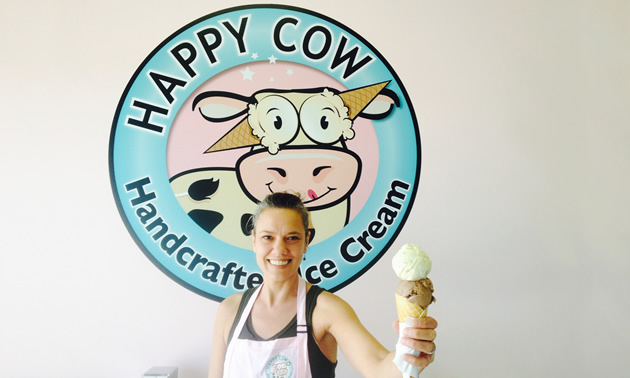 Co-owner Wendy Lyn holds out a two-scoop ice cream cone while standing in front of the Happy Cow Ice Cream's logo.