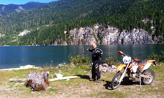 A man standing by a dirt bike and a lake. 