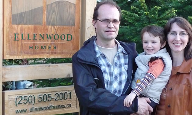 Man and woman standing together holding a young child in front of a wooden business sign