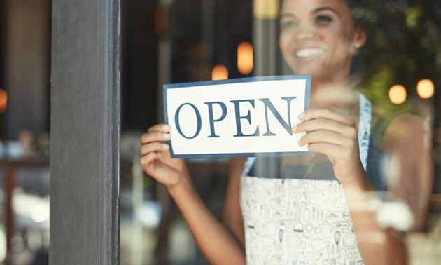 A woman is smiling behind a glass door, holding an OPEN sign up to the glass.