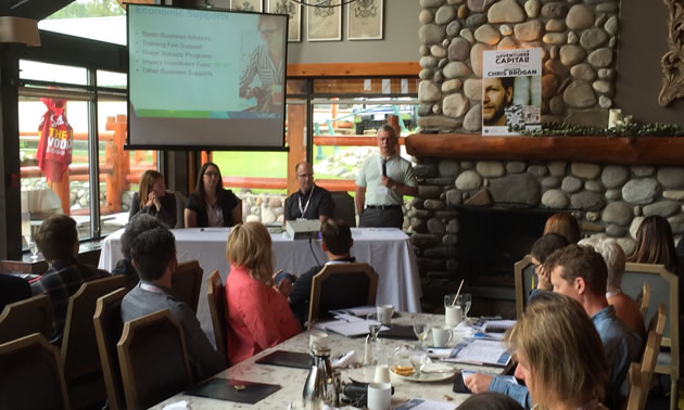 Group of people sitting at tables, watching a presentation on a screen at the front of the room.  