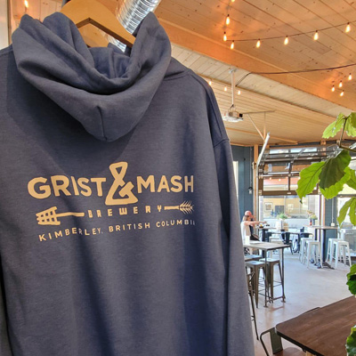 Grey Grist and Mash hoodie on hanger, out-of-focus interior shot of brewery on right-side of photo. 