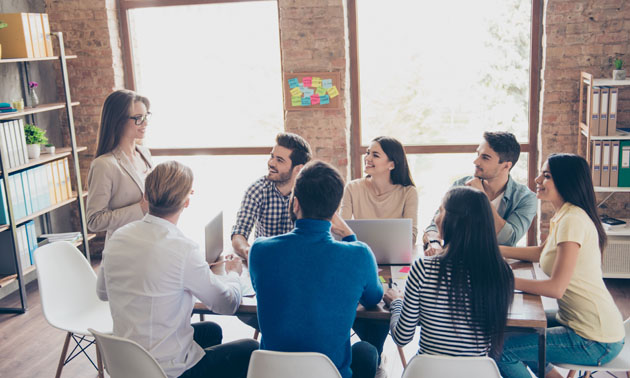 Group of people sitting around table in office setting, woman standing at head of table talking. 