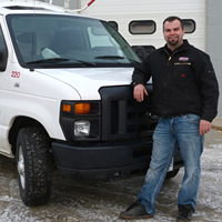 A man in a black jacket and blue jeans standing in front of a plumbing and heating van. 
