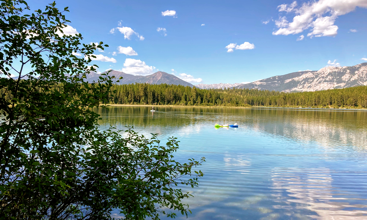 A canoer and people on floats enjoy a calm lake with mountains in the distance and a blue sky.