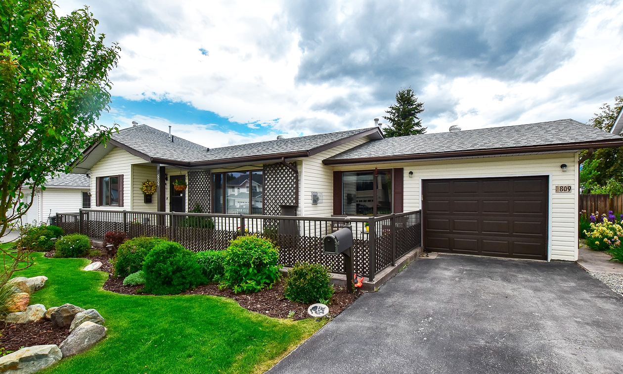 A one-storey tan and brown house with an attached garage has a well-maintained lawn and gardens with shrubs, trees and rocks.